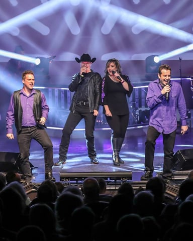 Live music performance on stage with six performers singing into microphones, purple and blue stage lighting, audience in foreground.
