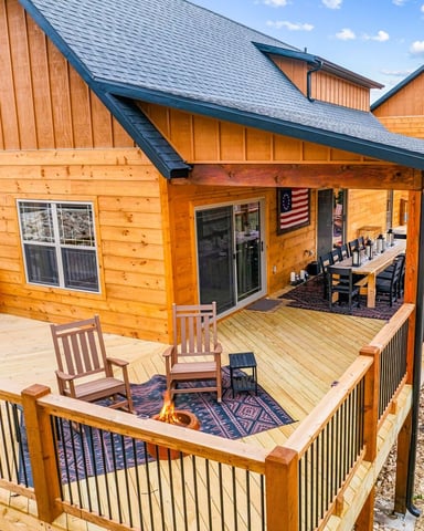 Modern log cabin with wraparound deck, fire pit, dining area, and American flag under blue-slate roof.