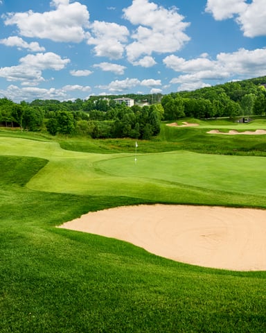 Golf course fairway with sand bunker, green grass, trees, and residential homes in distance under blue sky