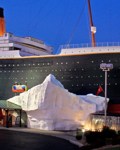 Titanic museum exterior at dusk with replica ship, large iceberg sculpture, and red flags illuminated against blue sky
