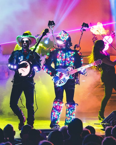 Live band performance with musicians wearing LED-lit costumes and instruments on stage with colorful lighting and smoke effects