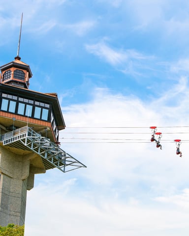 Tall observation tower with cupola and viewing platform, zip line riders suspended on cables against blue sky