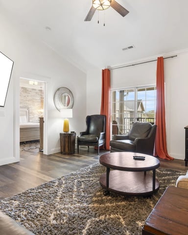 Bright living room with white brick fireplace, black leather chairs, beige sofa with orange pillows, round coffee table, and balcony access with orange