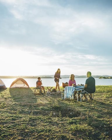 Family camping on grassy lakeside beach with two tents and folding chairs, lake and forested shoreline in background