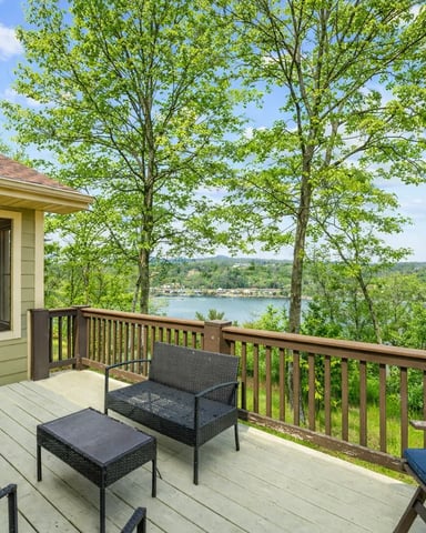 Deck with wicker seating overlooking forested hillside and lake with distant shoreline buildings