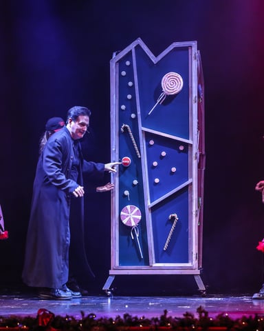 Theater stage performance with man in dark coat opening large blue cabinet filled with candy props, surrounded by children in colorful costumes