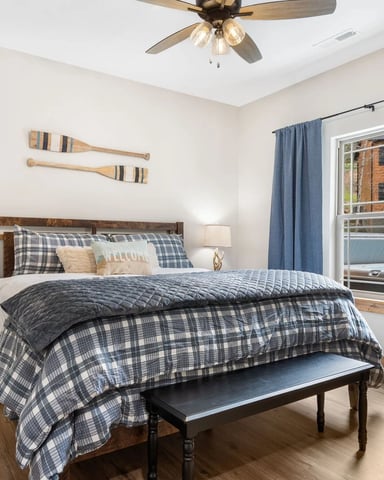 Bedroom with wooden bed, blue plaid bedding, nightstands with lamps, ceiling fan, and glass door to balcony overlooking brick building