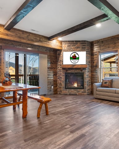 Spacious log cabin living room with stone fireplace, beige seating, wooden game table, and mountain views through glass doors.
