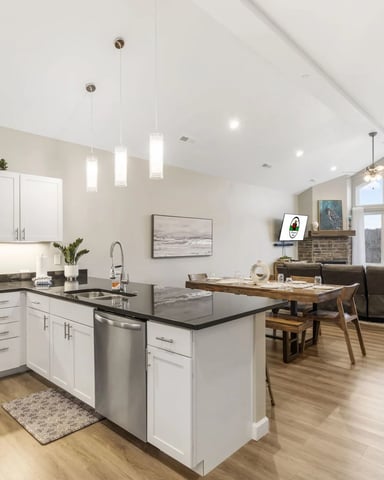 Open-plan kitchen and living room with white cabinetry, stainless steel appliances, black countertops, dining table, and brown sofa with large windows
