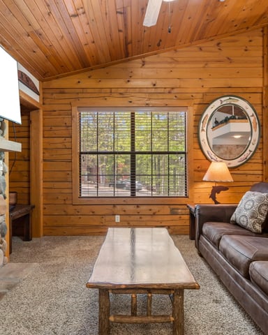 Rustic log cabin living room with stone fireplace, brown leather sofa, wooden beams, and large window overlooking forest.