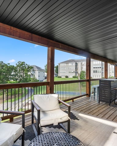 Covered deck with lounge chairs, dining table, and metal railing overlooking green lawn and residential buildings