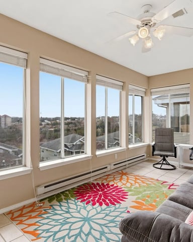Sunroom with expansive windows overlooking residential neighborhood, gray seating, colorful floral rug, and ceiling fan.