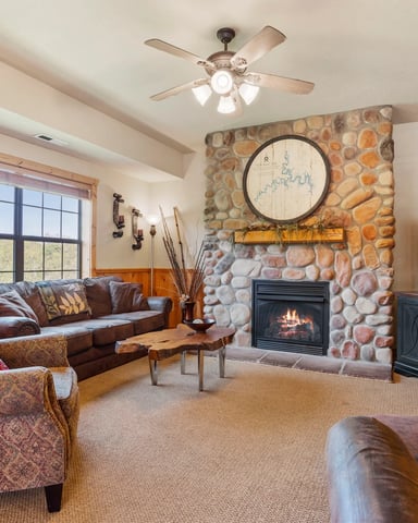 Spacious living room with stone fireplace, leather seating, TV, large windows, and ceiling fan