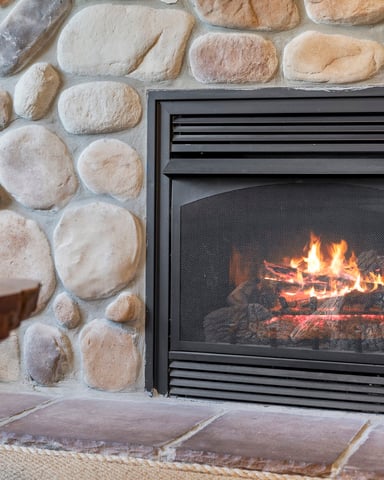 Gas fireplace insert with glowing flames built into tan stone wall, decorative bowl with dried grasses on side table
