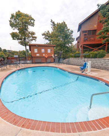 Outdoor swimming pool with brick deck, metal fence, and red cabin buildings surrounded by trees