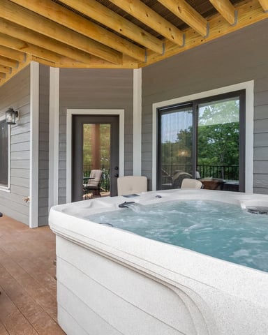 Covered patio with hot tub, lounge chairs, and glass doors opening to interior seating area with exposed wood beams