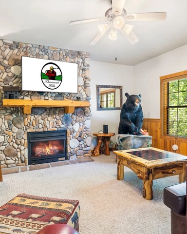 Rustic living room with stone fireplace, gas insert, wood beam mantel with TV, bear statue, pool table, and large windows overlooking trees