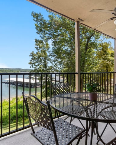 Covered lakeside patio with metal dining table, chairs, ceiling fan, and railing overlooking water and forested shoreline