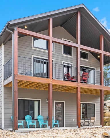 Modern two-story house with gray siding, red-brown wood frame, upper deck with red chairs, stone retaining wall, and turquoise Adirondack chairs on lower patio.