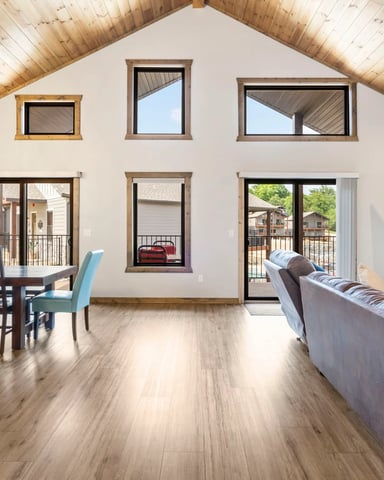 Open-plan living room with wood ceiling, dining table, sofa, fireplace, and multiple windows overlooking deck and surrounding buildings.