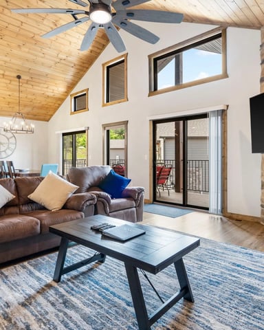 Open-plan living room with wood ceiling, brown leather sofa, stone fireplace, wall-mounted TV, and glass doors to deck