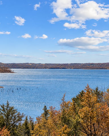 Large blue lake with autumn foliage along shoreline and forested hills under partly cloudy sky