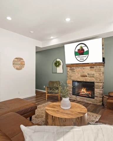 Living room with brown leather seating, wood coffee table, stone fireplace with active fire, sage green accent wall, and recessed lighting