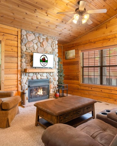 Rustic log cabin living room with stone fireplace, ceiling fan, brown leather seating, and windows overlooking green landscape.