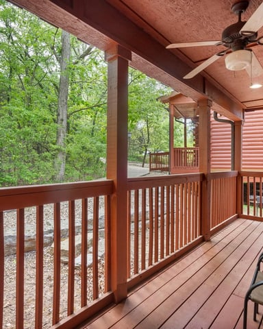 Covered wooden deck with ceiling fan, chairs, and forest views through railings