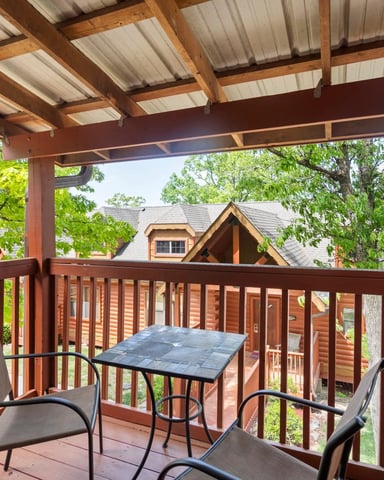 Covered deck with wooden railings, two chairs, small table, and view of neighboring log cabins surrounded by trees
