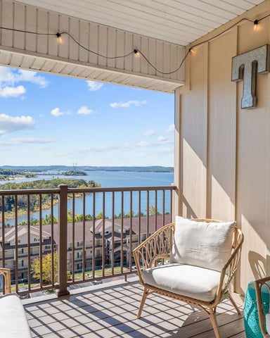 Covered balcony with wicker chairs overlooking lake, resort buildings, and forested shoreline with blue sky