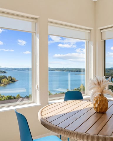 Dining room with large windows overlooking a blue lake, forested shoreline, and distant hills with blue chairs and wooden table