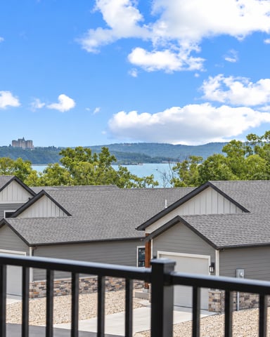 Modern townhouses with gray roofs overlooking a lake and forested hills with distant buildings, viewed from a black metal railing.