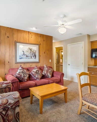 Open-plan living room with wood-paneled walls, red sofa, patterned armchairs, wooden coffee table, and kitchen with wood cabinetry visible in background.