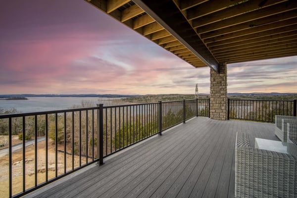 Covered deck with composite flooring, metal railings, and stone column overlooking lake and forested shoreline at sunset