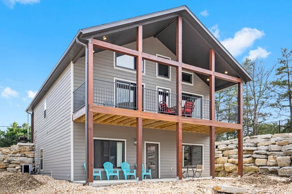 Modern two-story house with gray siding, red-brown timber frame, and upper deck with red Adirondack chairs overlooking wooded landscape.