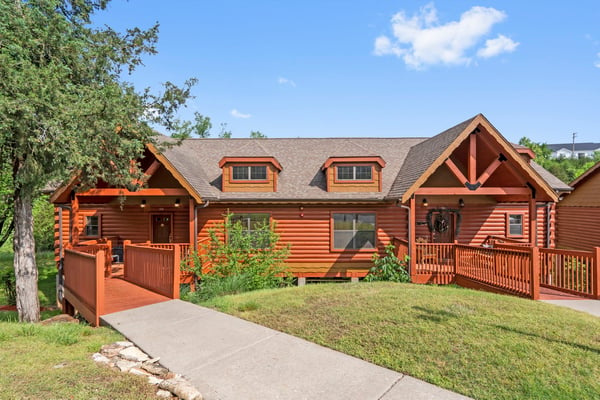 Large red log cabin with two dormers, wraparound decks, and concrete walkway on grassy lot with mature trees
