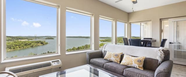 Bright living room with gray sofa, glass coffee table, and expansive windows overlooking a lake and green shoreline