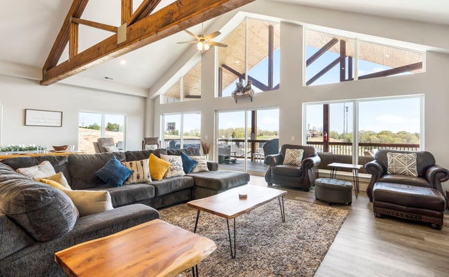 Open-plan living room with exposed wood beams, dark leather sectional, and floor-to-ceiling windows overlooking a lake and deck.