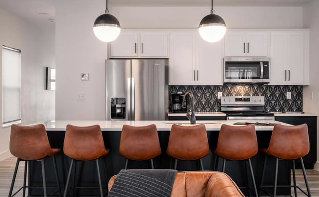 Modern kitchen with white cabinetry, stainless steel appliances, black geometric backsplash, and black island with six cognac leather bar stools