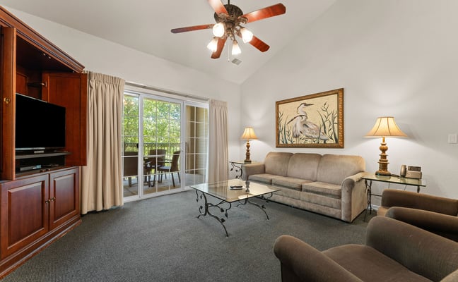 Spacious living room with beige sofa, wood entertainment center, glass doors to balcony, ceiling fan, and framed heron artwork.