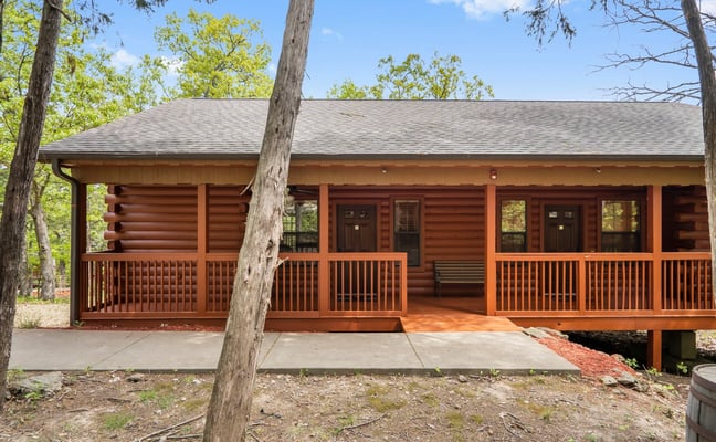 Single-story log cabin with wraparound deck and carport, surrounded by tall trees