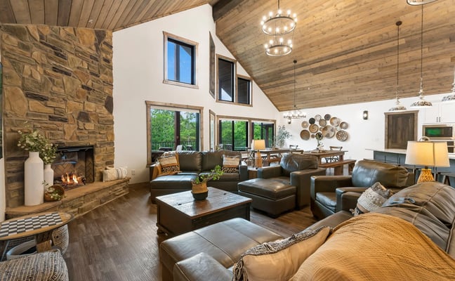 Open-plan living room with stone fireplace, leather seating, wood ceiling, and forest views through glass doors