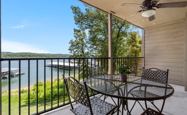 Covered lakeside patio with metal dining table, chairs, ceiling fan, and railing overlooking water and forested shoreline