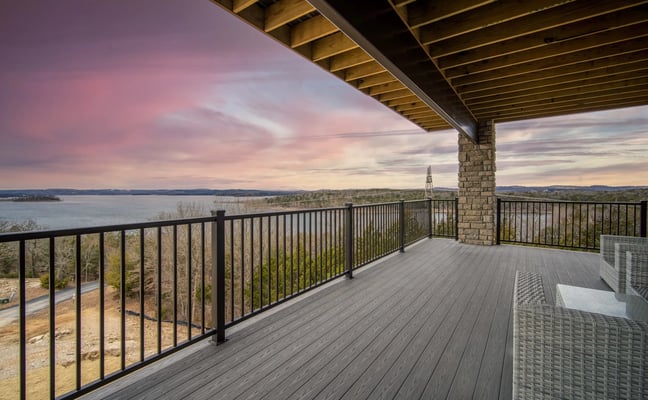 Covered deck with composite flooring, black metal railing, and stone column overlooking lake and forested shoreline at sunset